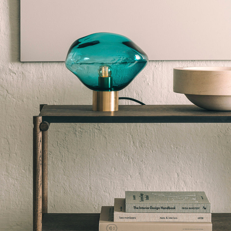 Crystal Stone bordlampe i fargen blågrønn fra Hadeland Glassverk. Bordlampen vises med messing fot på et skatoll.