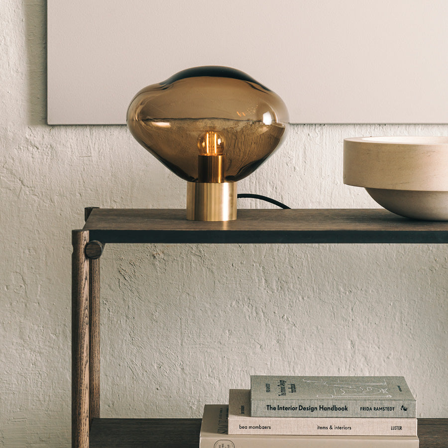 Crystal Stone bordlampe i fargen kakao fra Hadeland Glassverk. Bordlampen vises med messing fot på et skatoll.
