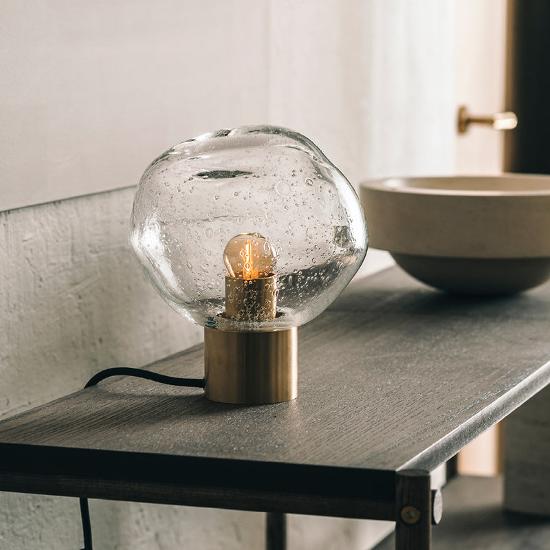 Crystal Stone bordlampe i klar soda fra Hadeland Glassverk. Bordlampen vises med messing fot på et skatoll.