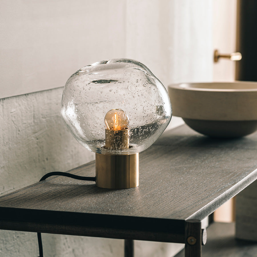 Crystal Stone bordlampe i klar soda fra Hadeland Glassverk. Bordlampen vises med messing fot på et skatoll.