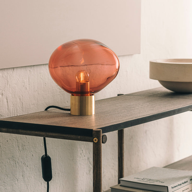 Crystal Stone bordlampe i fargen morgenrød fra Hadeland Glassverk. Bordlampen vises med messing fot på et skatoll.