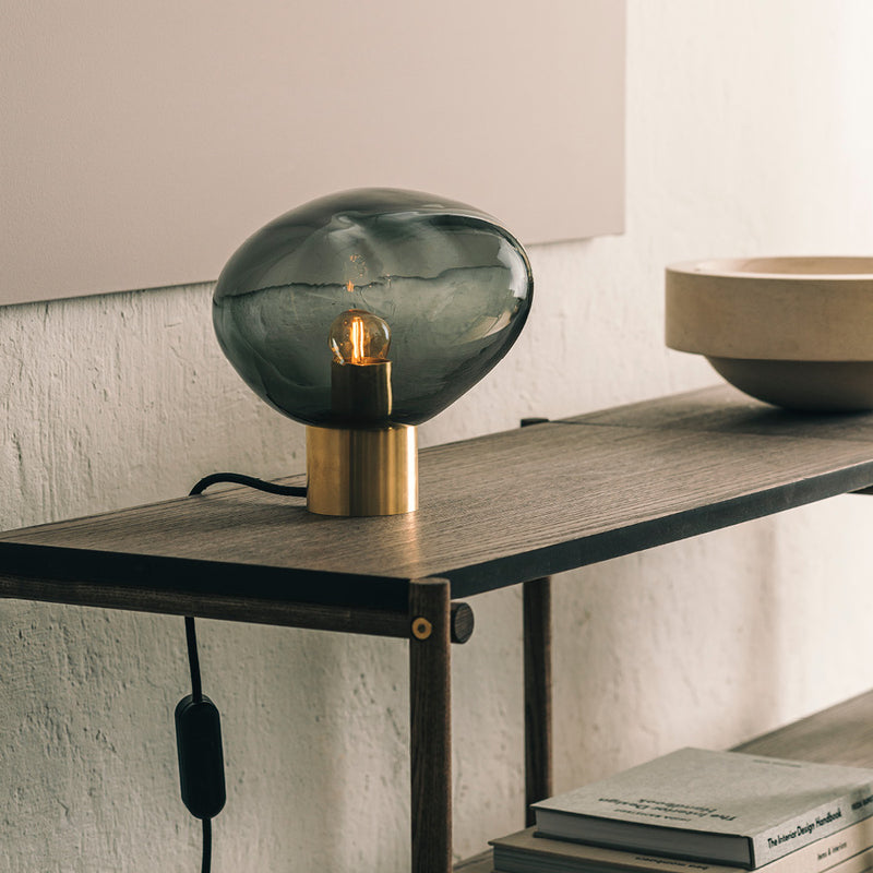 Crystal Stone bordlampe i fargen røkgrå fra Hadeland Glassverk. Bordlampen vises med messing fot på et skatoll.